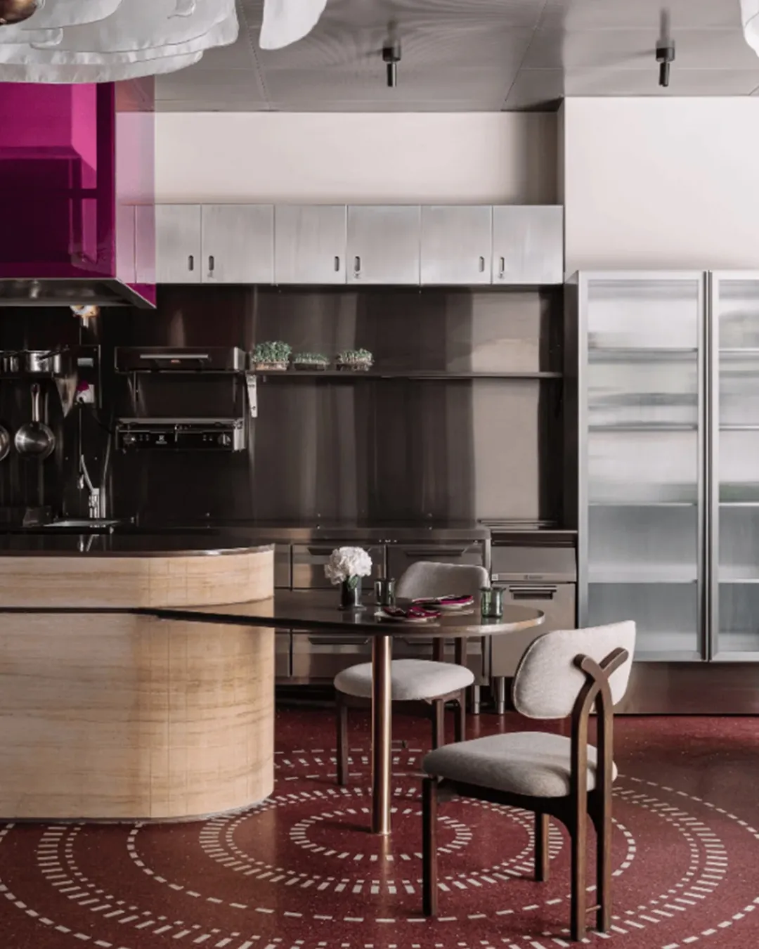 Open restaurant kitchen with stainless-steel cabinets, pink-tinted glass hood detail, and a round dining table featuring minimalist décor, set against red circular-pattern flooring.