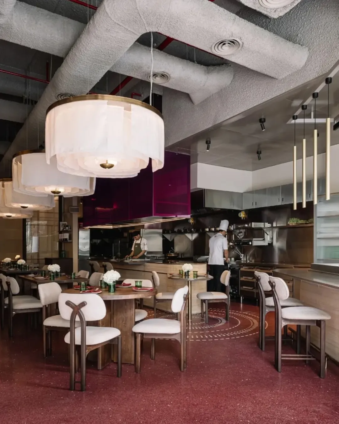 Contemporary restaurant dining area with luxurious wooden tables, soft neutral chairs, and dramatic overhead lighting. The open kitchen is visible in the background.