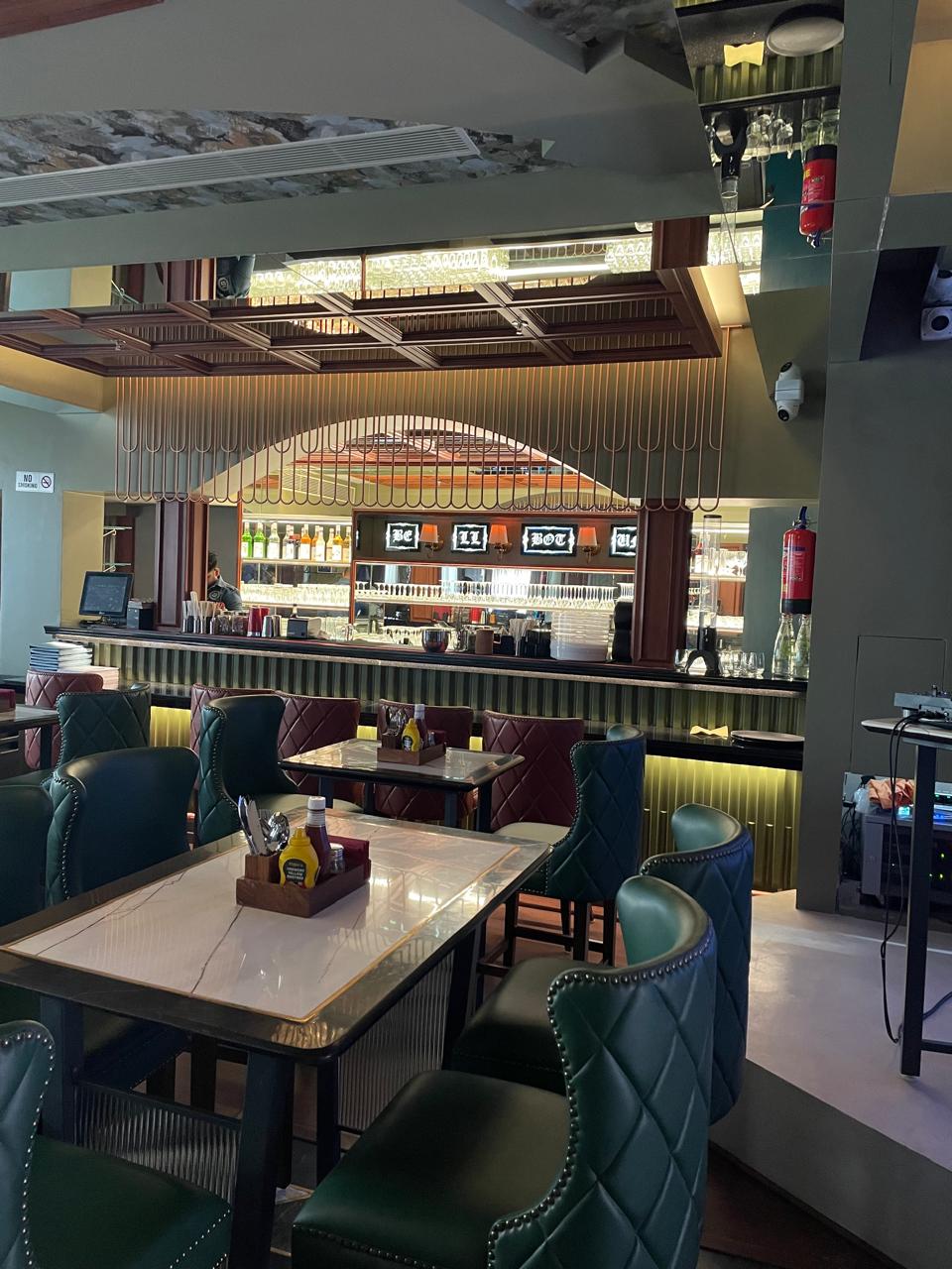 Modern restaurant interior with a bar, shelves of bottles, and green and burgundy leather chairs around marble tables. The space has a stylish, contemporary design.