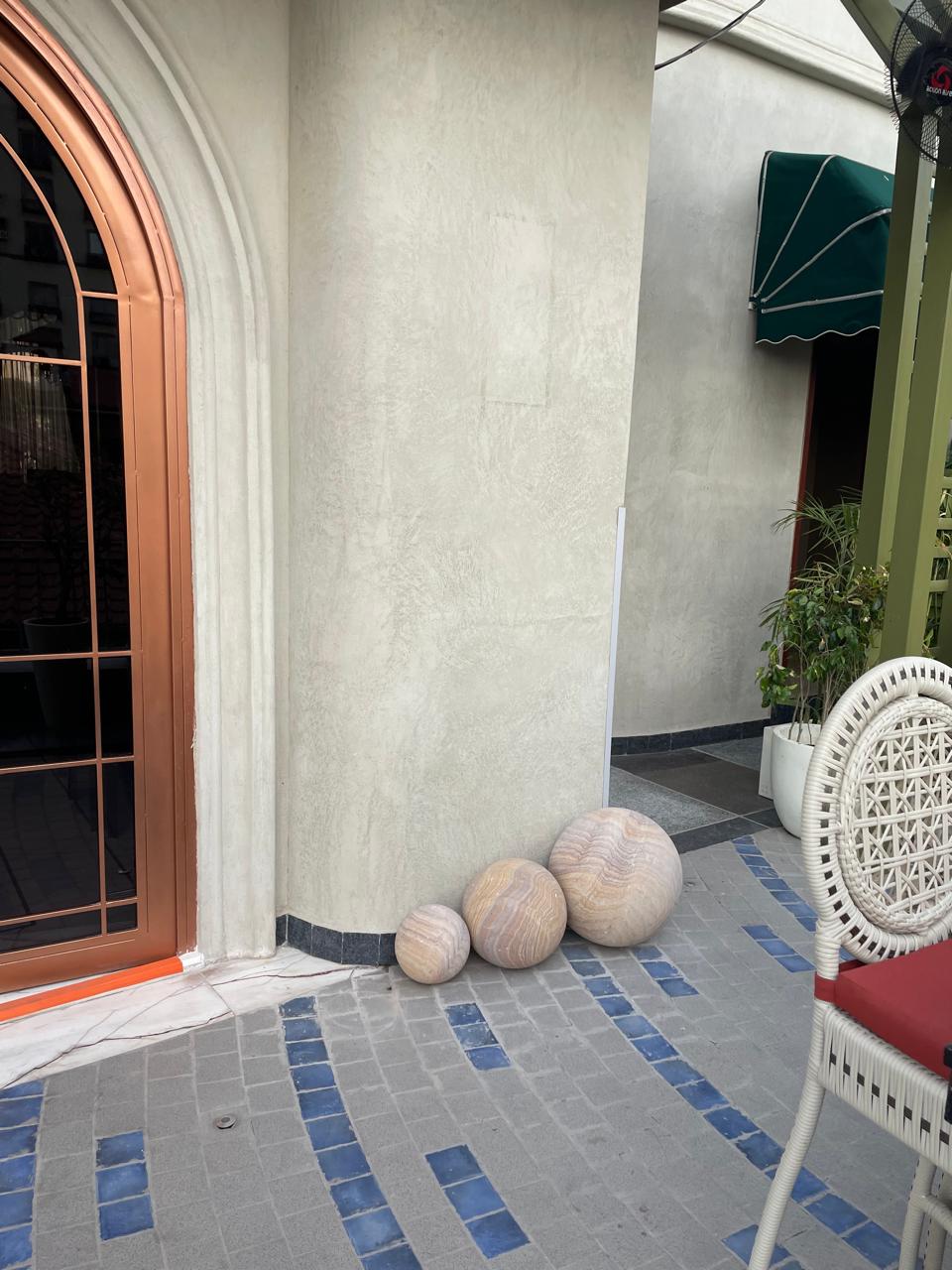 Decorative stone spheres placed near a copper-framed door on a patterned tiled floor with blue and gray stones. The image features a minimalist exterior with a green canopy above a potted plant, adding a touch of modern design to the architectural space.