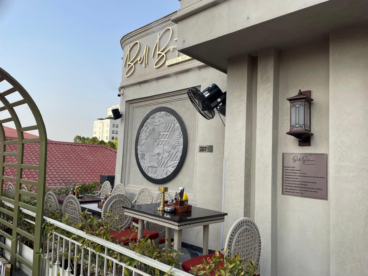 Outdoor rooftop dining area of Bell Bar featuring elegant woven chairs, black tabletops, decorative wall art, and warm ambient lighting, with a view of red-tiled rooftops and nearby buildings under a clear sky.