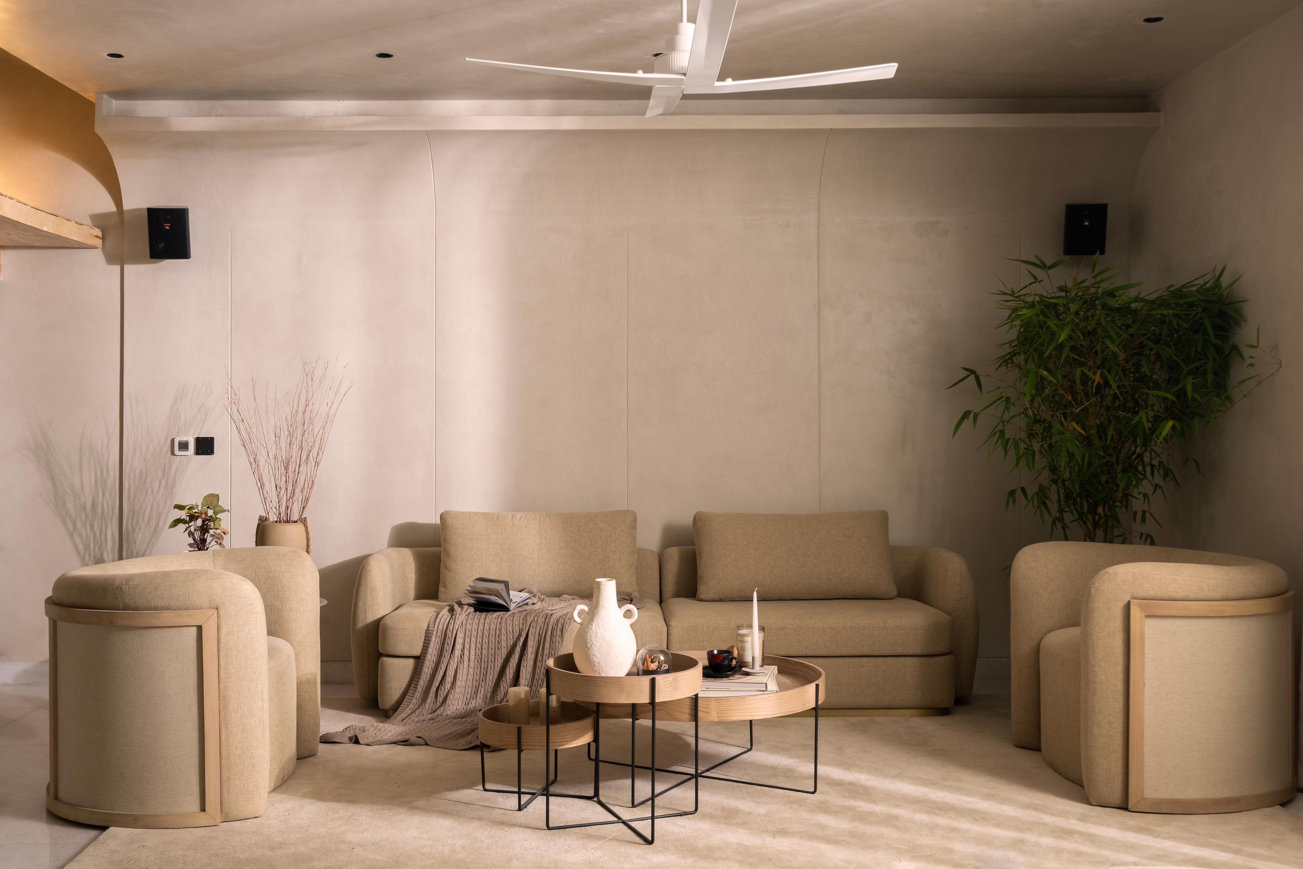 Cozy living room with beige sofas, soft chairs, and a wooden coffee table. There’s a plant, a vase, candles, and a blanket, making the space look calm and inviting.