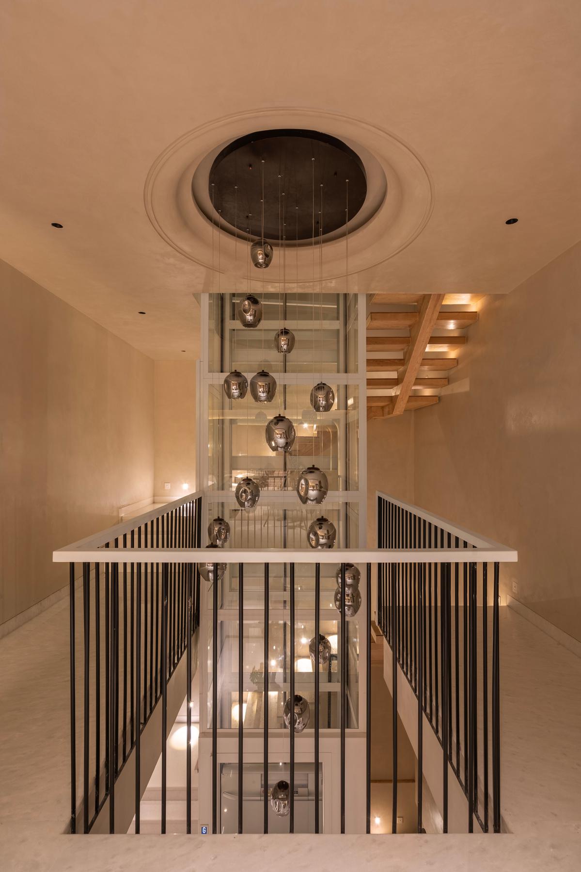 Modern hallway with a glass elevator, hanging glass lights, and a wooden staircase. The space has beige walls, black railings, and warm lighting for a clean, contemporary look.