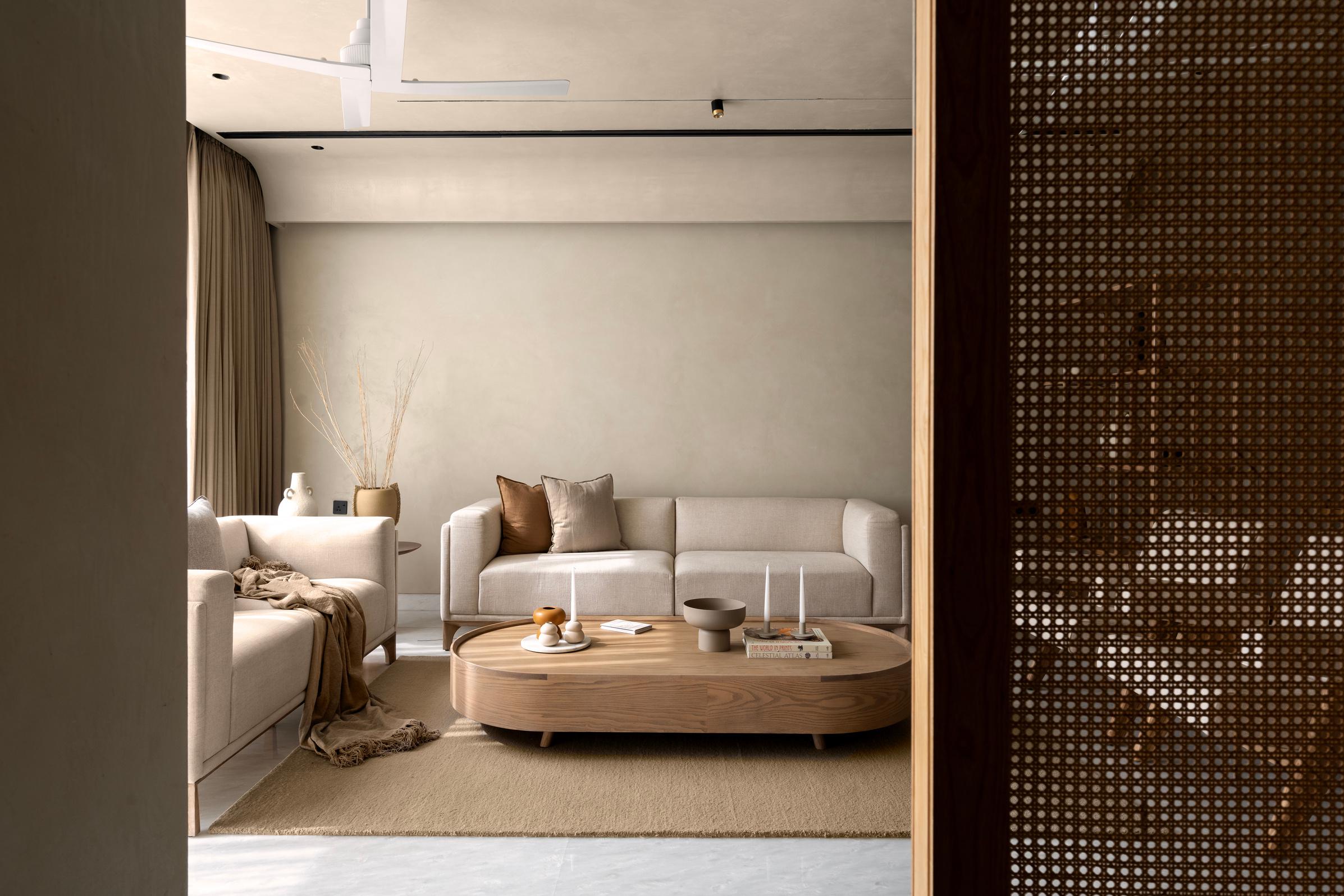 Elegant and serene living room with a minimalist design. The beige sectional sofa is complemented by a round wooden coffee table, decorated with candles, decorative bowls, and books. Soft, neutral-colored throws and cushions add texture to the space. The room features a natural woven rug, a large vase with dried branches, and floor-to-ceiling curtains. A modern ceiling fan and a wooden partition add to the contemporary style of the space.