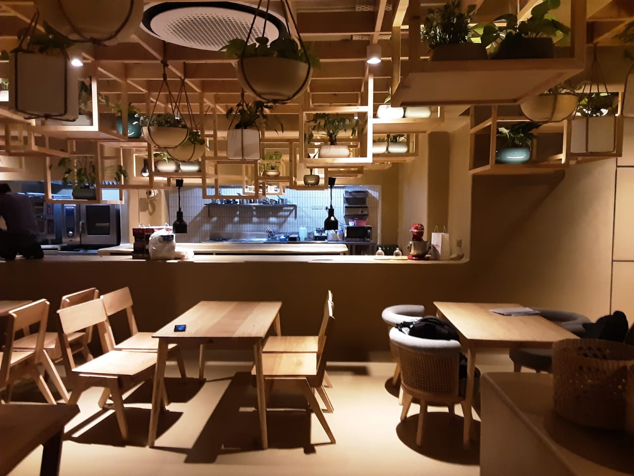 Modern minimalist café interior with wooden tables and chairs, geometric hanging planters, and a visible kitchen area with cooking appliances and soft lighting.