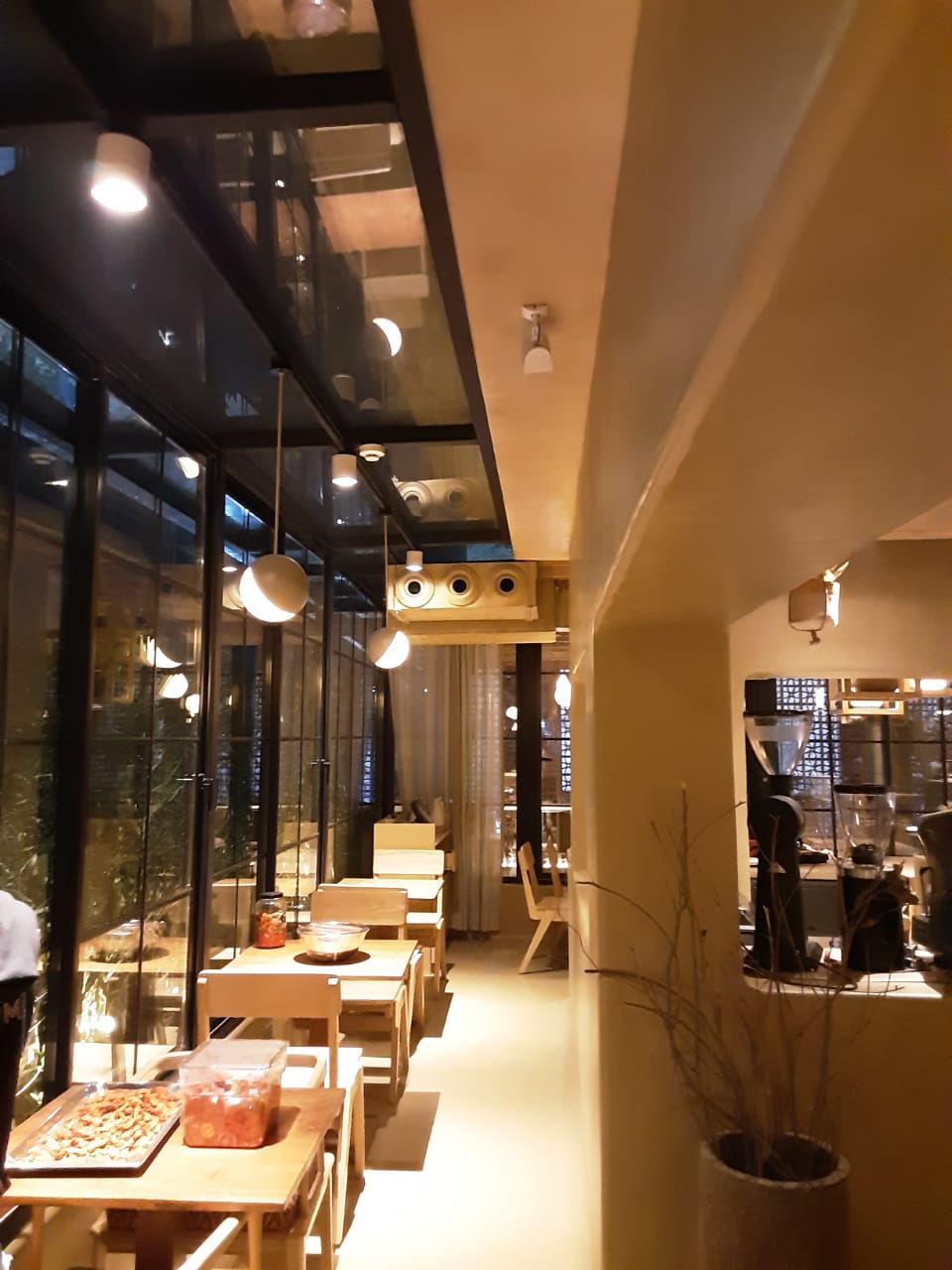 Interior of a modern café with a minimalist design, showcasing a narrow hallway lined with wooden tables. Above, the ceiling features a glass panel and spherical light fixtures, creating a bright, open atmosphere. The space is illuminated with soft lighting and has large windows offering an outdoor view.