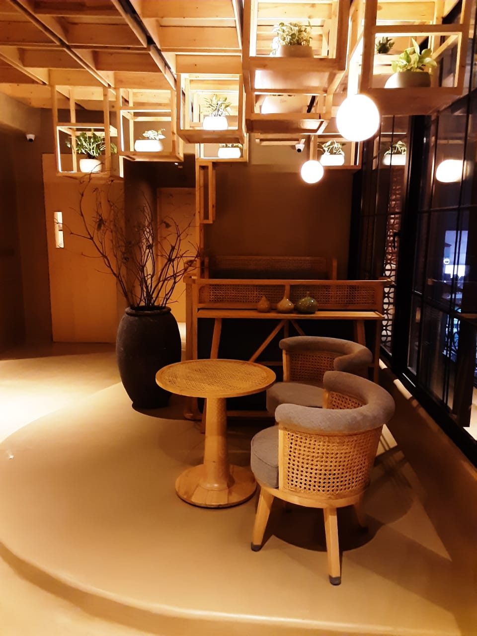 Modern café corner with minimalistic wooden furniture, featuring a round table and two rattan chairs with gray cushions. The ceiling has stylish geometric wooden light fixtures with hanging plants and soft lighting, creating a cozy, serene atmosphere.