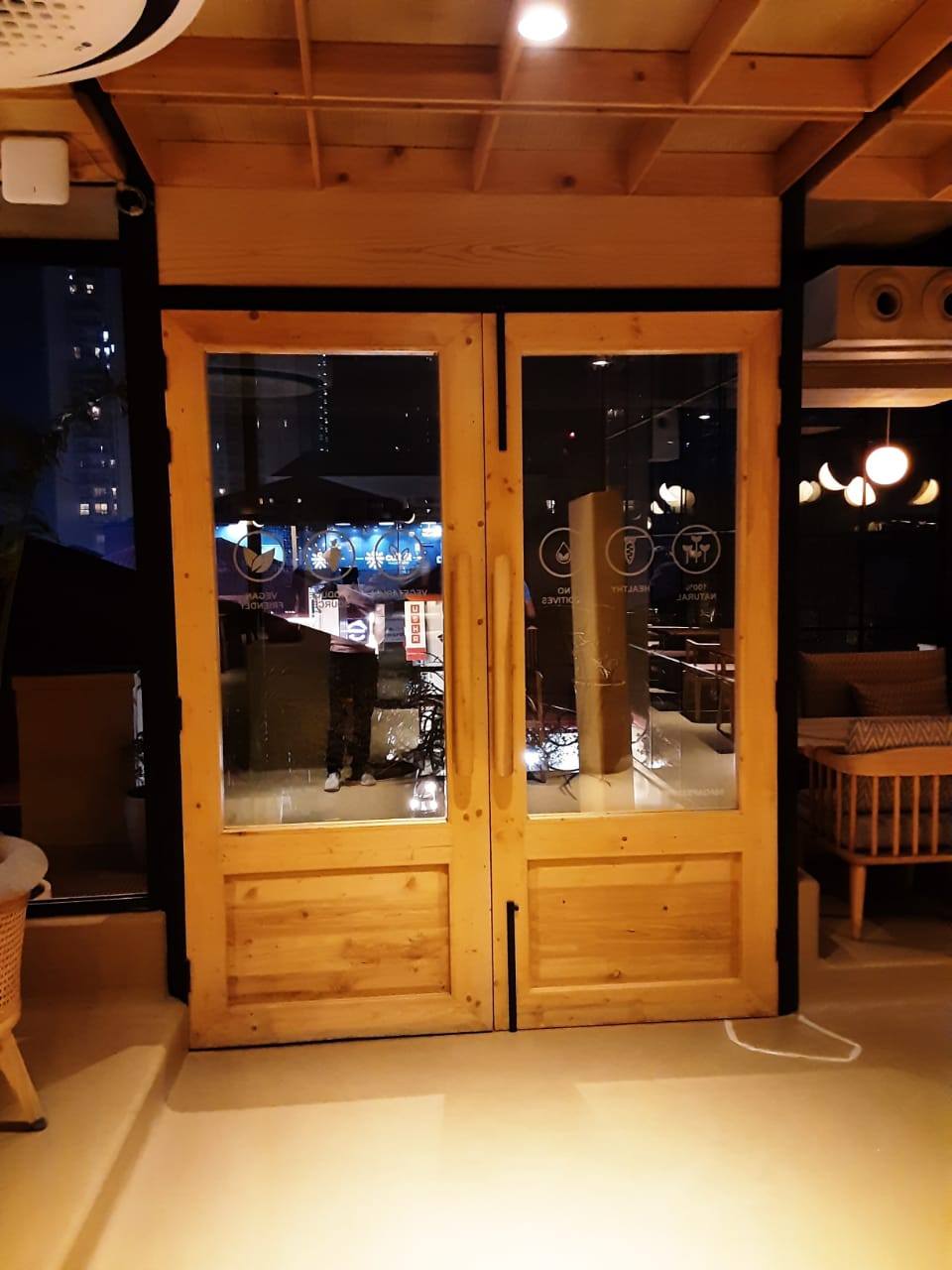 Wooden double doors with large glass windows reflecting the interior of a modern café, showcasing dim lighting, contemporary furniture, and a cozy ambiance. A person is visible in the reflection.