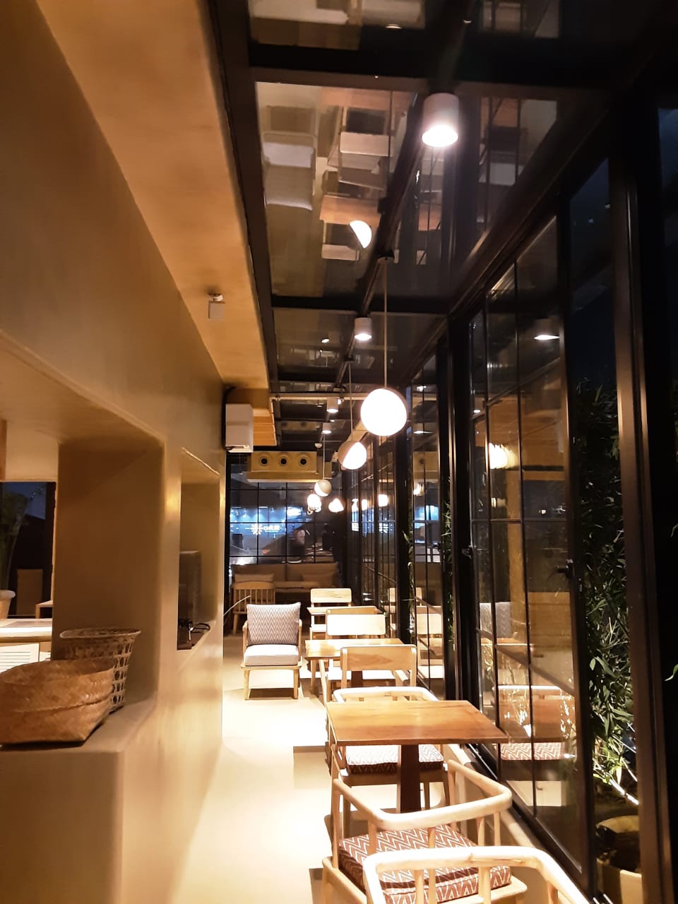 Cozy glass-walled café corridor at night with a row of wooden tables and cushioned chairs, warm globe pendant lights and mirrored ceiling reflections creating an intimate, ambient dining space.