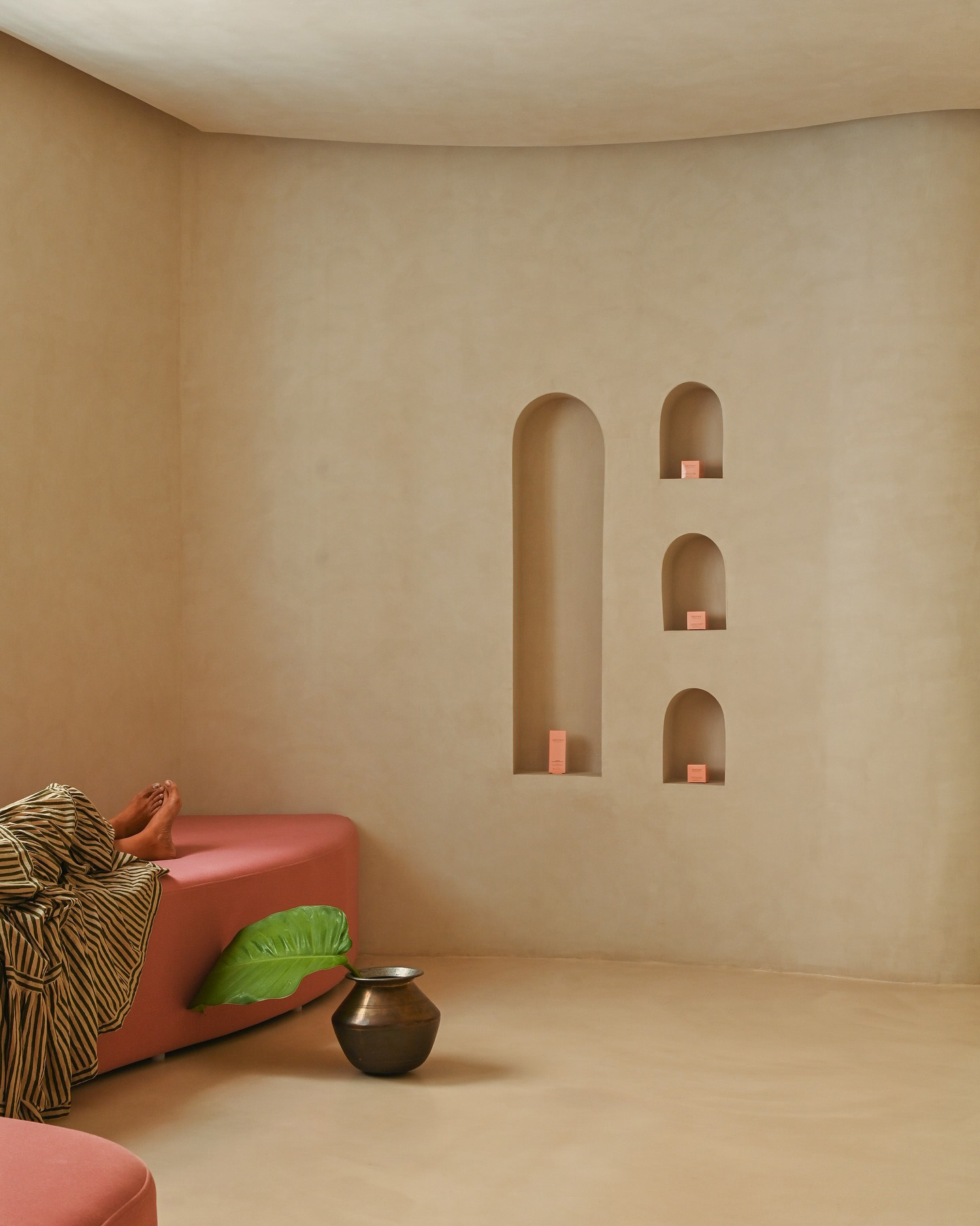 Bare feet and striped fabric draped over a curved pink bench in a minimalist cream-toned interior, with recessed arched wall niches holding small peach-colored objects, a bronze vessel on the floor and a large green leaf.