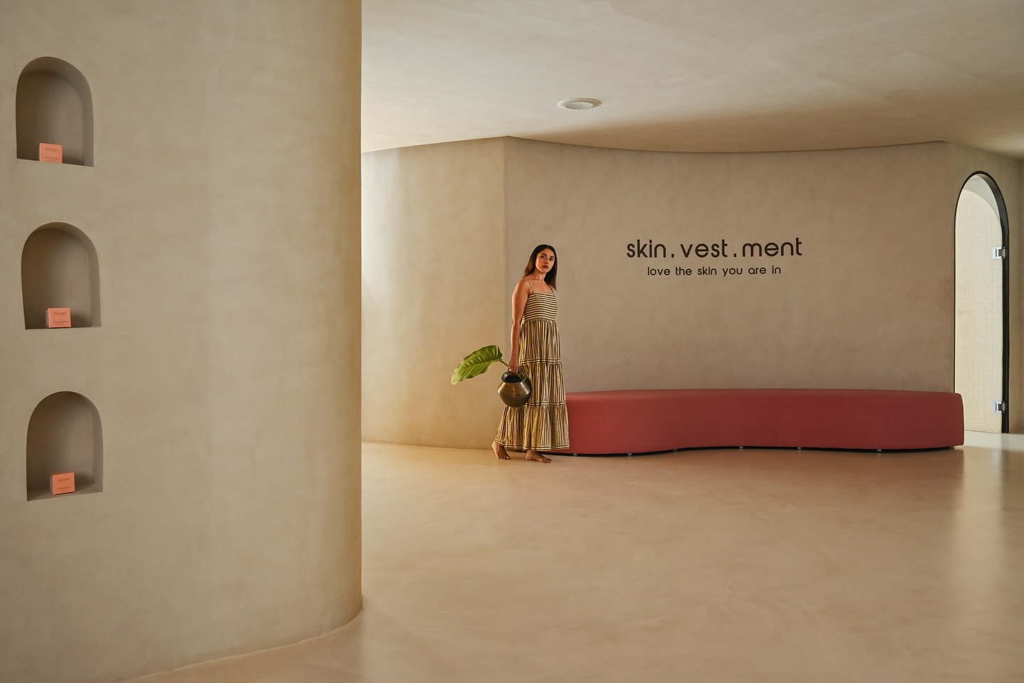 Woman in a striped maxi dress standing in a minimalist, cream-toned interior beside a curved pink bench and “skin.vest.ment” wall signage, with recessed niches displaying peach-colored product boxes.