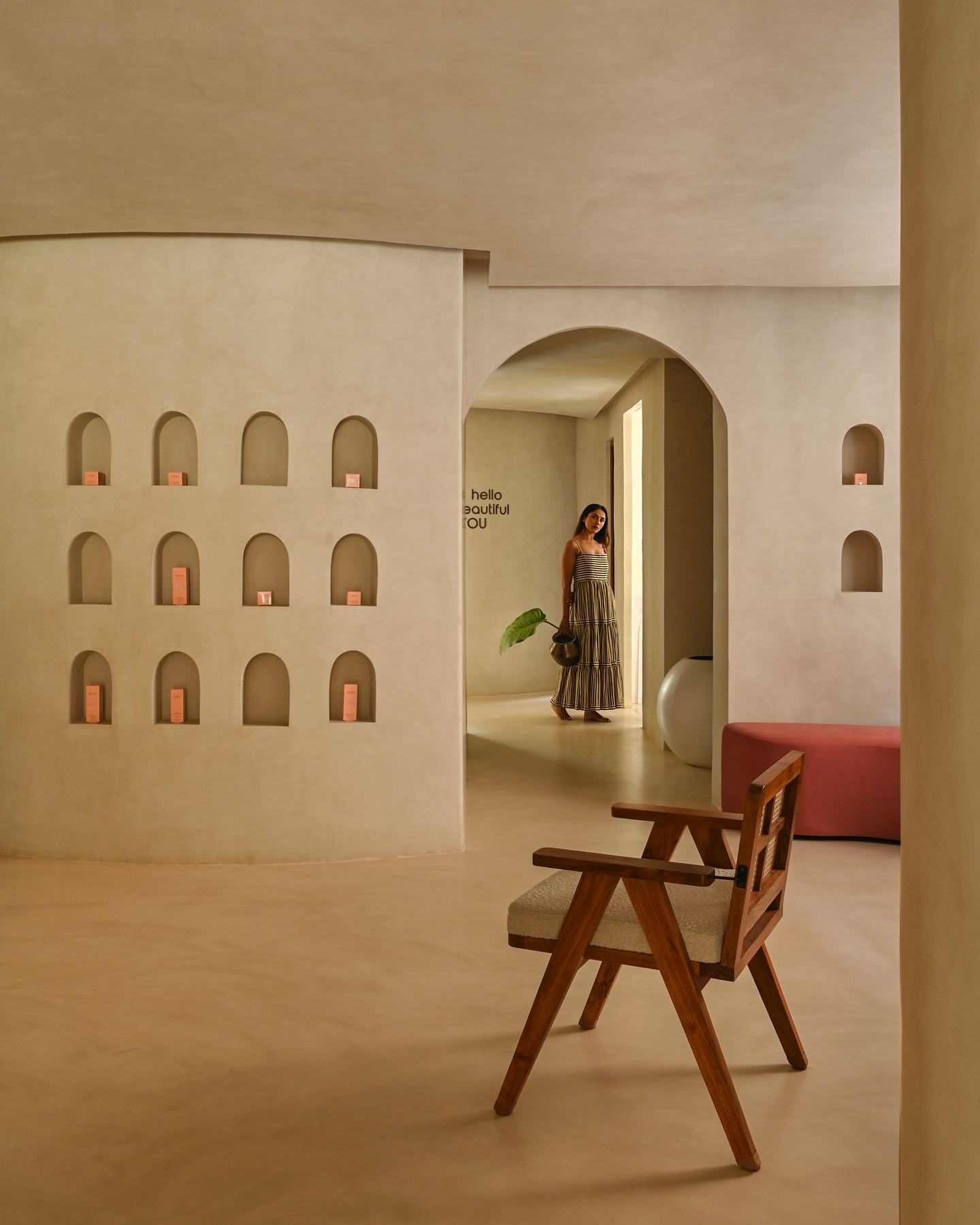Woman standing in a sunlit minimalist cream interior with an arched doorway, recessed product niches, and a wooden armchair in the foreground.