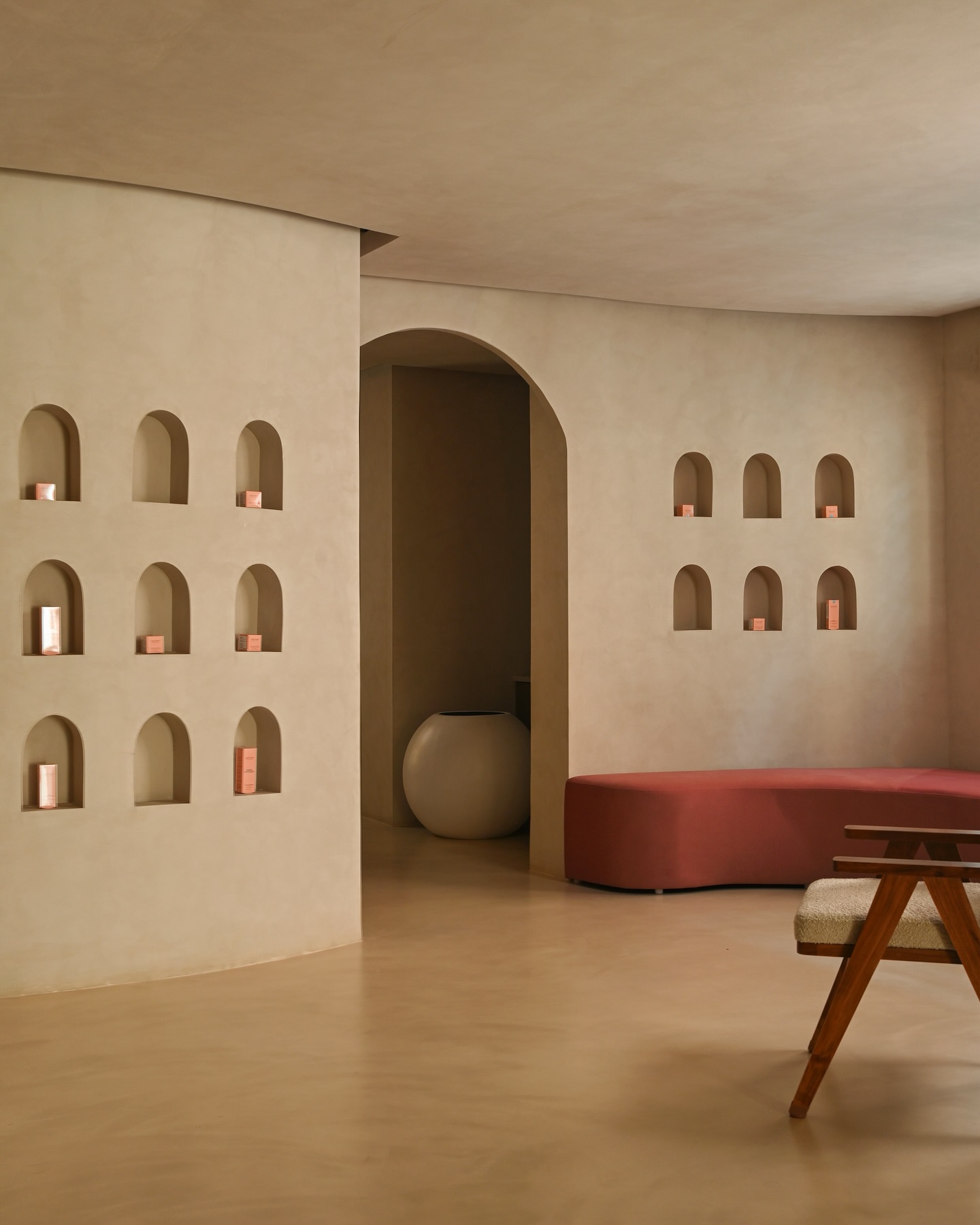 Minimalist gallery lounge with curved beige plaster walls, rows of small arched niches holding product boxes, an arched doorway, and a curved red bench.
