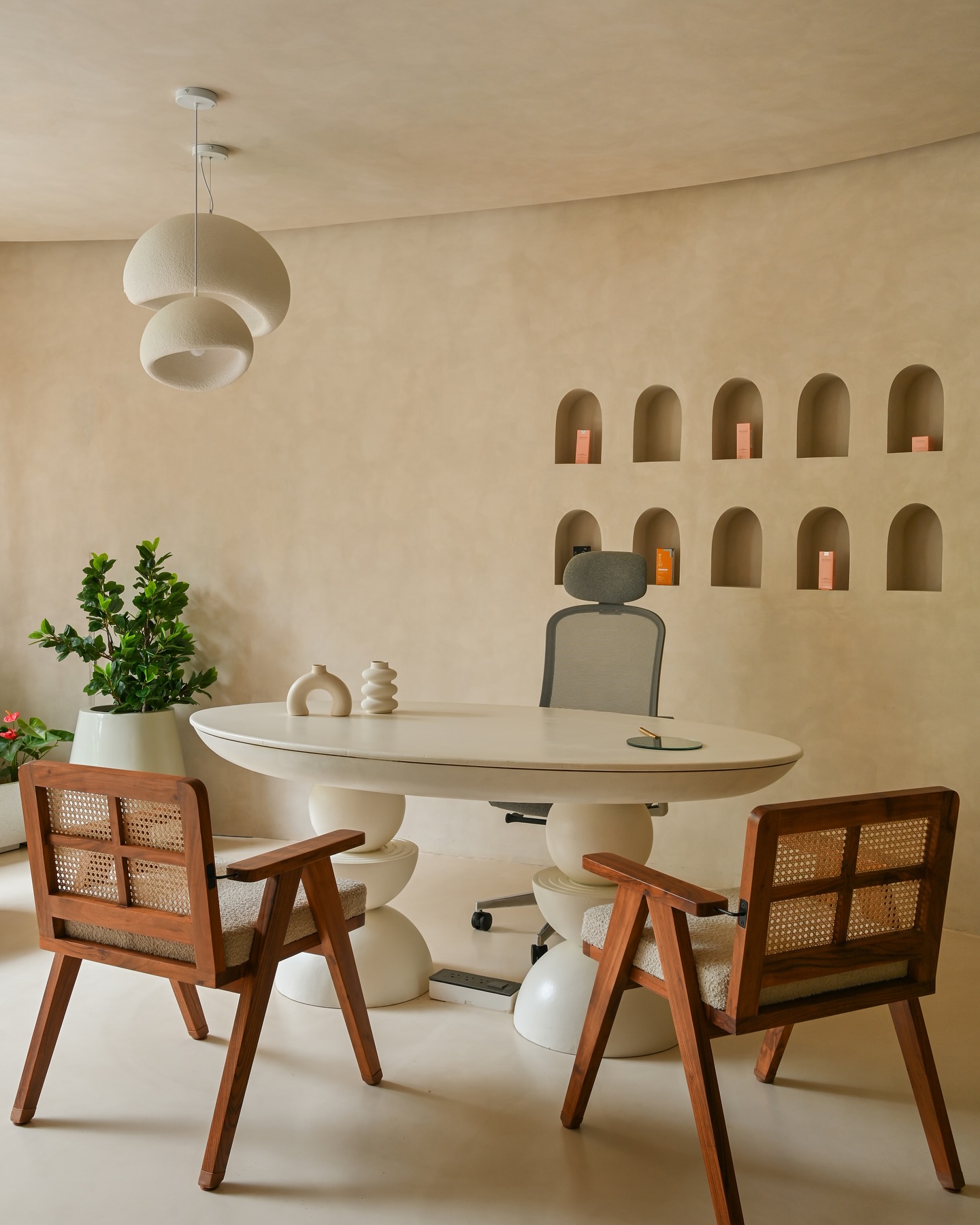 Modern gallery meeting room with a sculptural white oval table, wooden cane-back chairs, curved beige walls, built-in arched niches, and indoor greenery