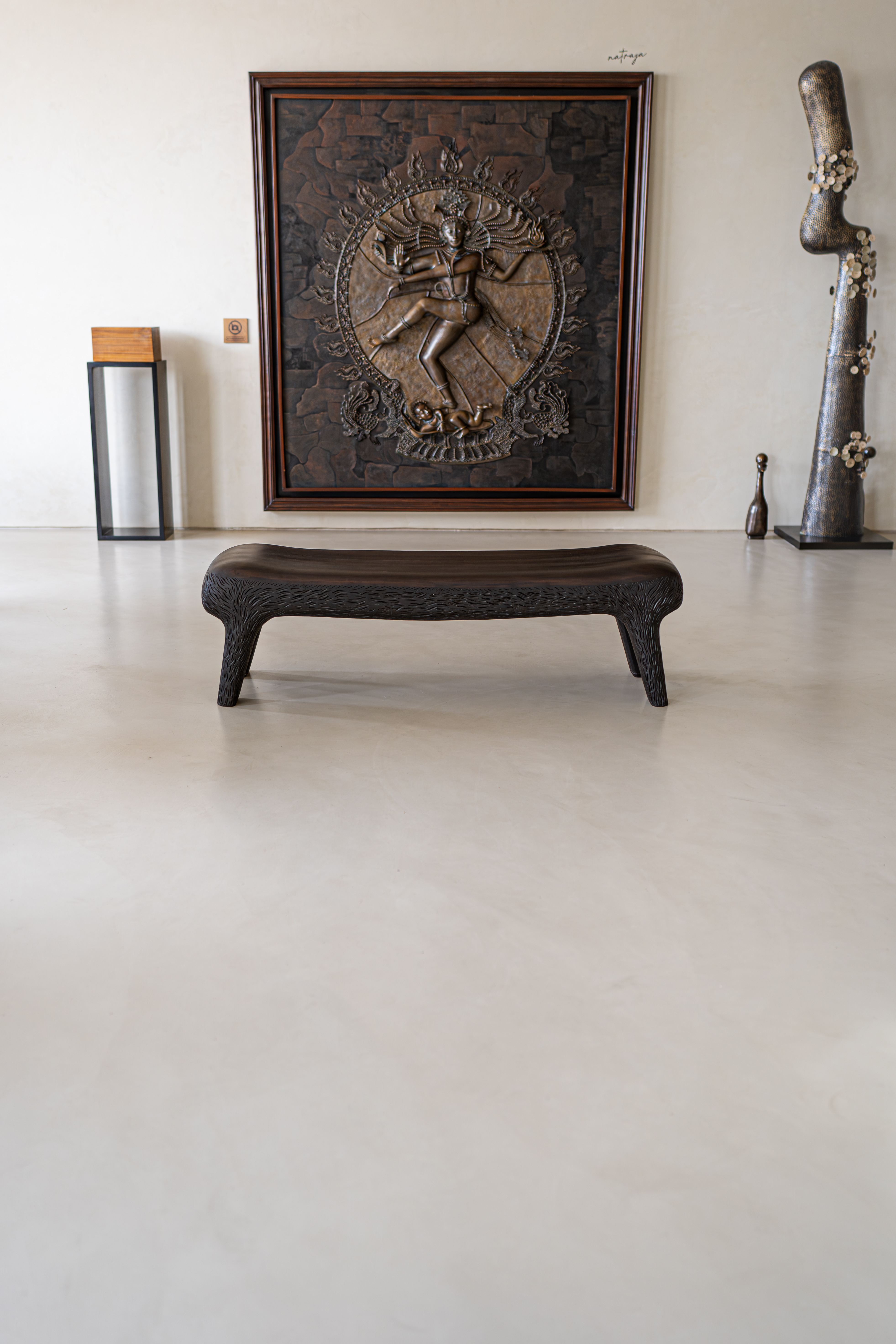 Art gallery interior featuring a large bronze Nataraja wall sculpture, a carved dark wooden bench, and a tall textured metal sculpture on a polished light floor.