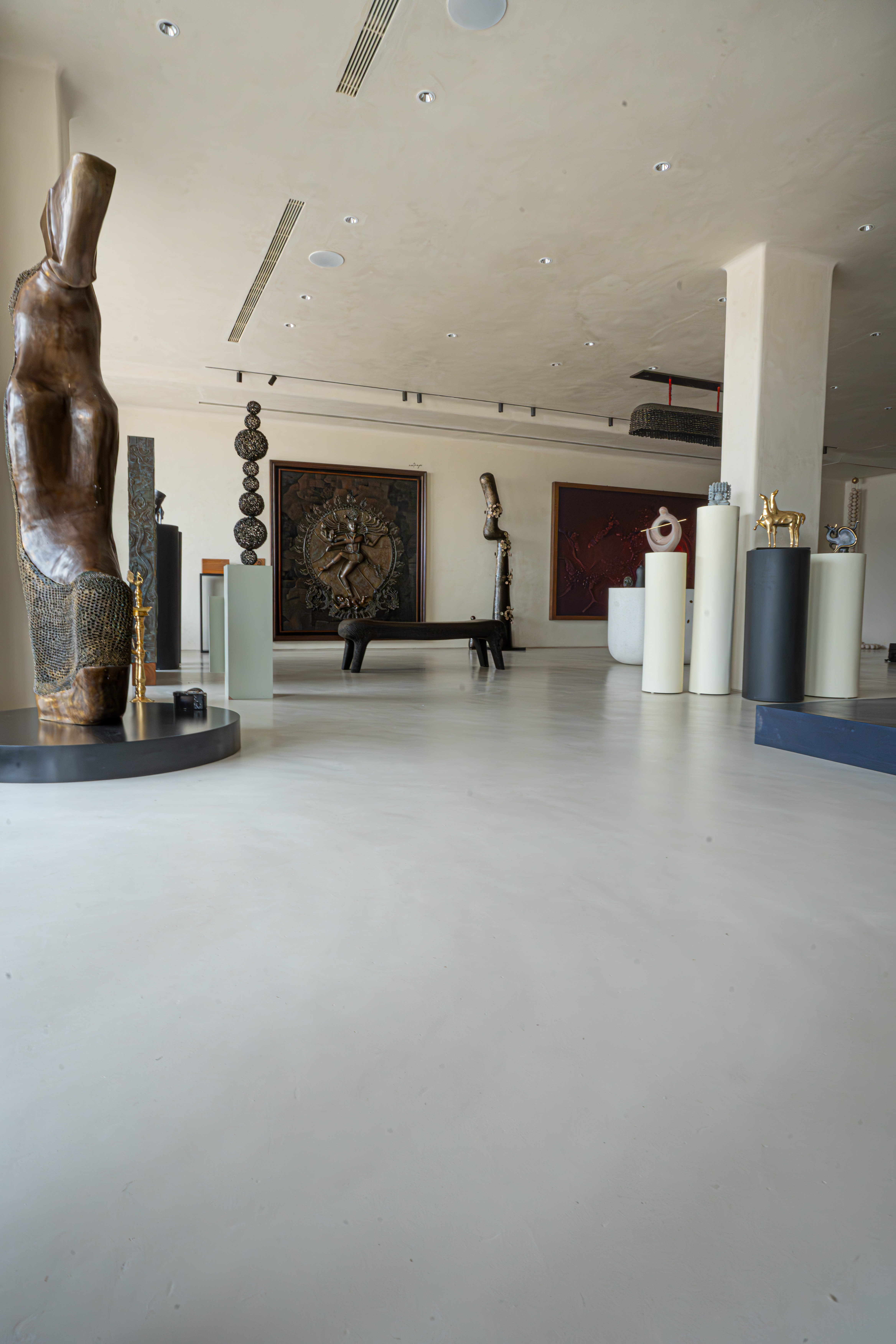 Contemporary art gallery interior with a tall bronze torso on a black plinth, pedestals displaying gold figurines, and a red spiral bas-relief on the back wall.