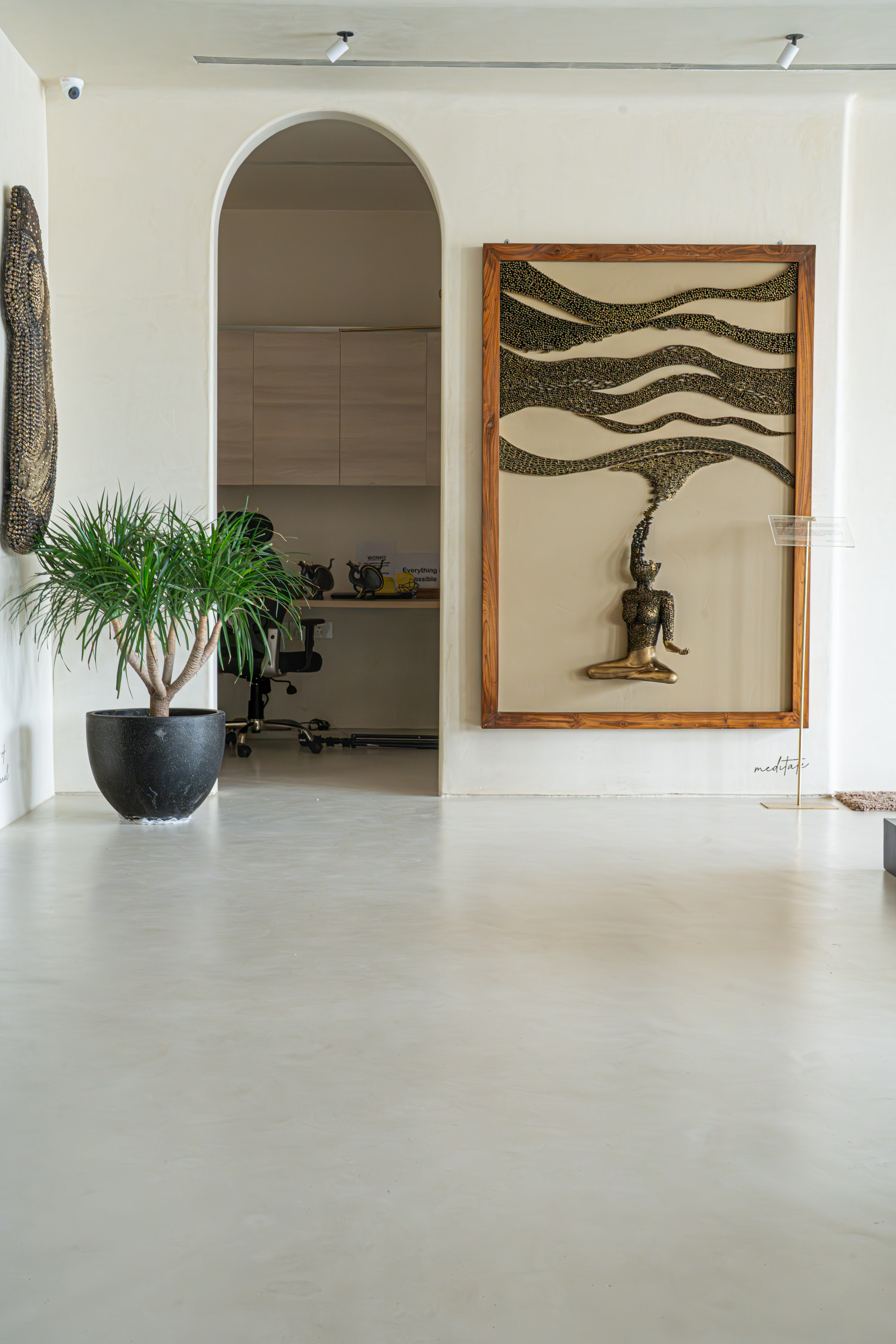 Minimalist art gallery entrance interior featuring an arched doorway, large framed brass meditation wall sculpture with cascading beadwork, a black planter holding a ponytail palm, and a pale polished concrete floor.